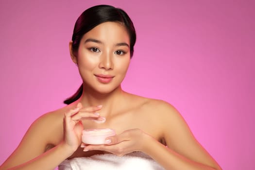 A young woman holds a skincare cream tub in her hands while posing against a vibrant pink background. Soft lighting highlights her radiant complexion and confident expression.