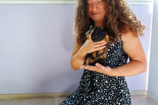 A woman wearing a patterned dress sits comfortably while holding her guinea pig gently in her hands. The relaxed atmosphere is enhanced by soft natural light.