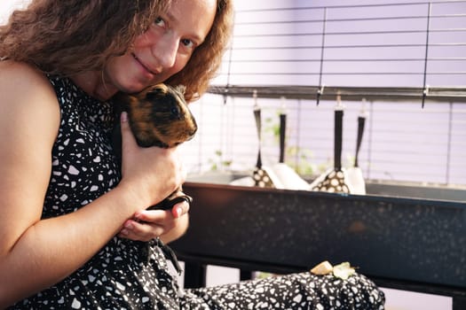 A young girl smiles as she gently cradles a guinea pig close to her chest. She sits comfortably beside a pet cage, creating a warm atmosphere for bonding with her furry friend.