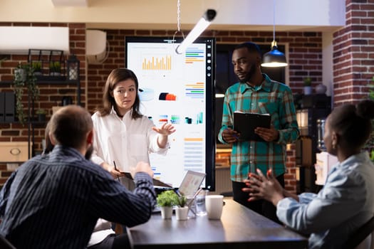 Male and female business executives stand in front of LED monitor, presenting financial data and project plans in brick wall workspace. Diverse entrepreneurs reviewing marketing trends on screen.