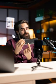 Tech guru informing audience about newly released mini soundbar. Man in studio pressing buttons on high end music playing device, testing specifications, filming with smartphone