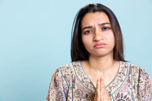Religious indian girl praying to god and showing optimism on camera, talking to jesus with respect and devotion against blue background. Christian faithful woman expresses her belief, worship.