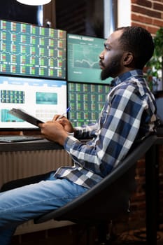Focused stockbroker with clipboard, studying forex market trends at night, using multiple monitors in modern office. Black man reviewing financial data and stock charts displayed on computer screens.