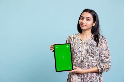 Indian girl smiling and showing isolated copy space screen in front of camera, creating an advertisement for a commercial against blue background. Young joyful adult holding device in studio.