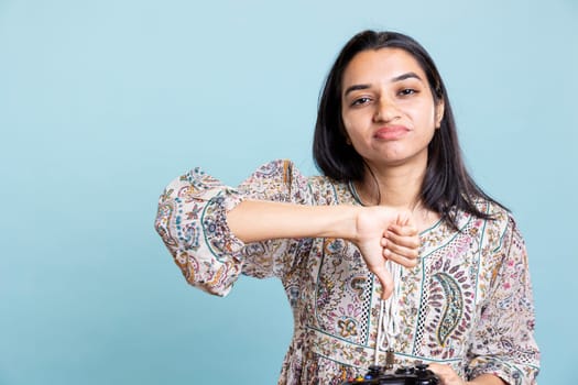 Indian displeased girl feels disappointed after losing video game, giving dislike sign against blue background. Irritated frustrated woman lost gaming tournament, controller in hand.