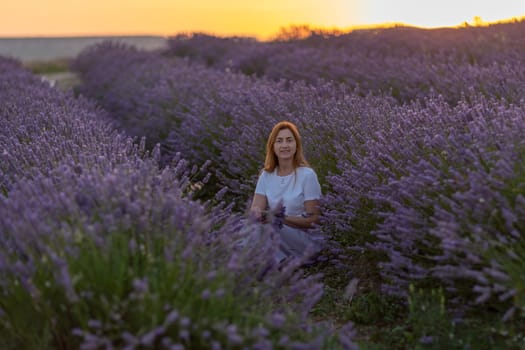 A woman sits in a field of lavender flowers. The flowers are purple and the sky is orange
