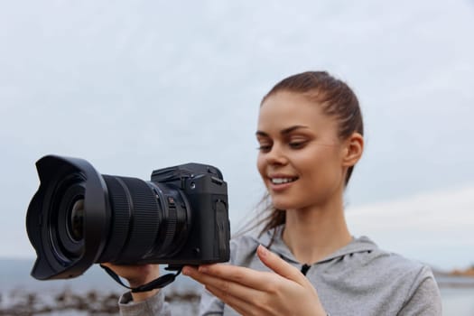 A cheerful young woman wearing casual gray clothing holds a camera at the beach, surrounded by a calm ocean landscape Capturing the essence of creativity and passion for photography