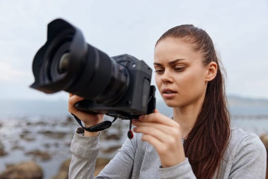 A young woman with long hair focused on her camera, dressed in casual grey attire by the sea, radiating passion for photography with a tranquil seascape