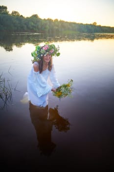 Slavic plump chubby girl in long white dress on the feast of Ivan Kupala with flowers and water in a river or lake on summer evening