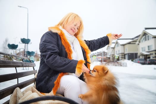 A middle-aged woman with blonde hair enjoys a winter walk, playfully interacting with her fluffy Pomeranian in the snow.