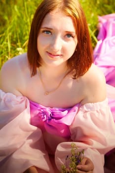 A young woman sits in lush grass, wearing a flowing pink dress, enjoying a sunny day while holding delicate flowers.