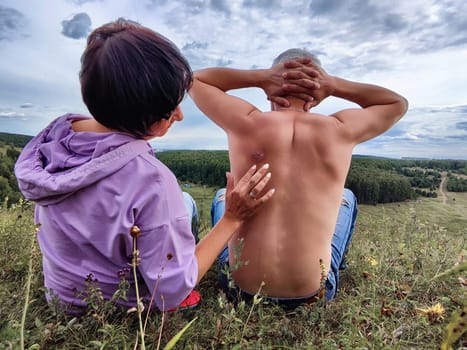 Two people relax on a hillside, appreciating nature while one gently touches the other's back, surrounded by greenery.