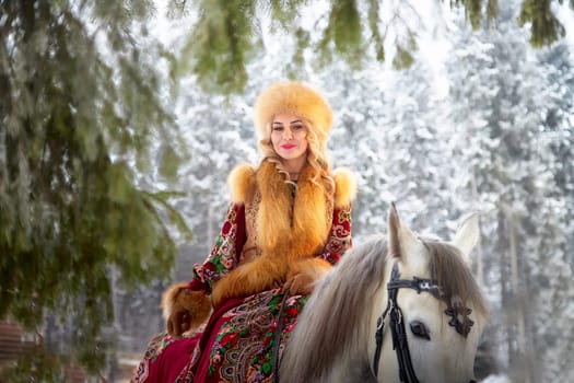 A beautiful Russian girl in a vibrant national dress poses on a horse during the winter celebration of Maslenitsa.