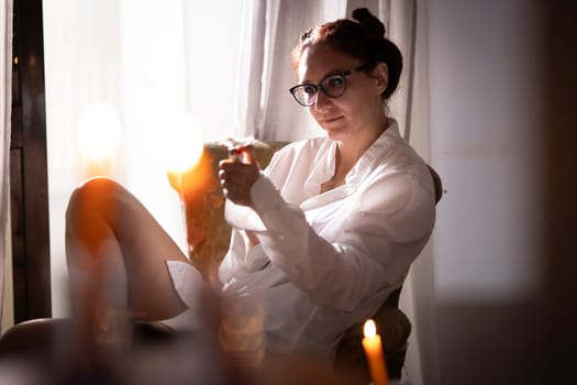 Serene woman in white shirt enjoying a quiet moment at home, lighting a candle by the window in warm sunlight