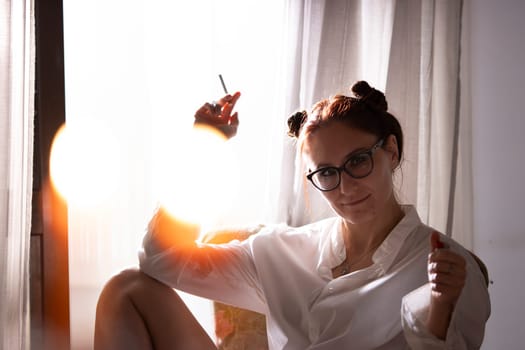 Relaxed woman vaping and looking at camera near window with bright backlight