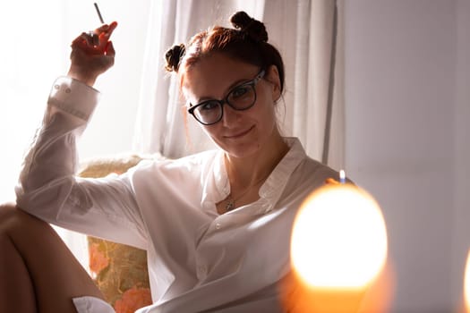 Redhead woman wearing glasses and white shirt smoking a cigarette, sitting on an armchair near a window with candles in the foreground