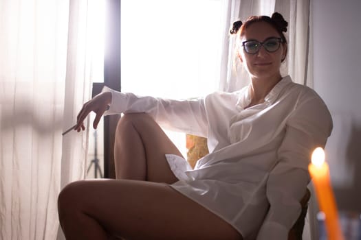 Young woman wearing glasses and white shirt relaxing at home and smoking a cigarette by the window