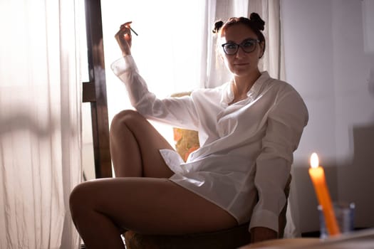 Woman wearing eyeglasses and white shirt sitting near window holding pen with candlelight in foreground