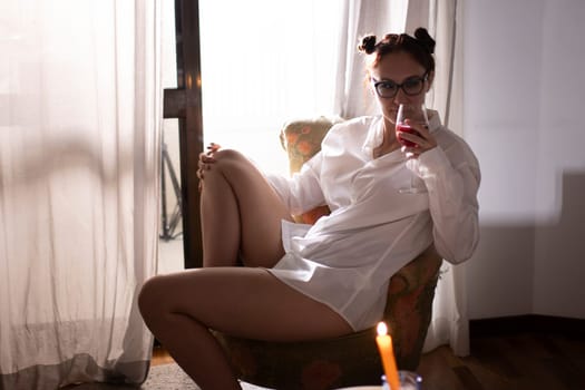 Young woman wearing a white shirt enjoying a glass of red wine by candlelight in an armchair