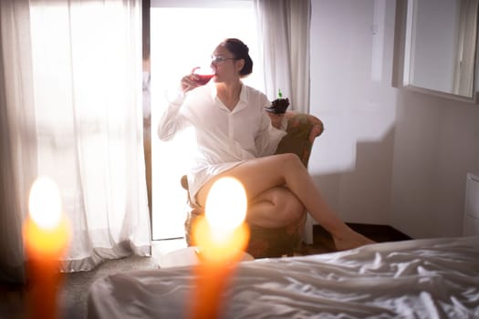 Woman enjoying wine and cake while sitting in armchair near window in hotel room