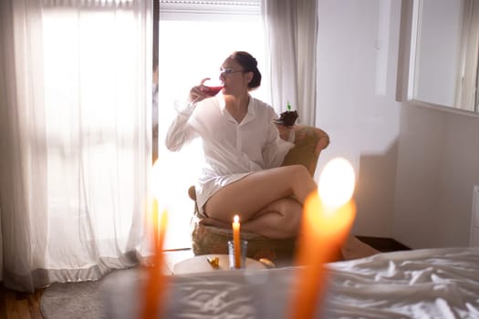 Woman wearing a white shirt sitting in an armchair drinking wine and eating a slice of cake with a candle on it