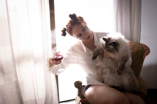 Young woman holding a glass of wine while cuddling her cat, sitting on a vintage armchair by a bright window