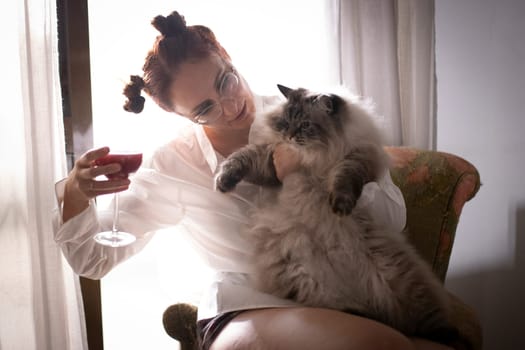 Young woman enjoying a glass of red wine while holding her fluffy cat, relaxing by the window in natural light