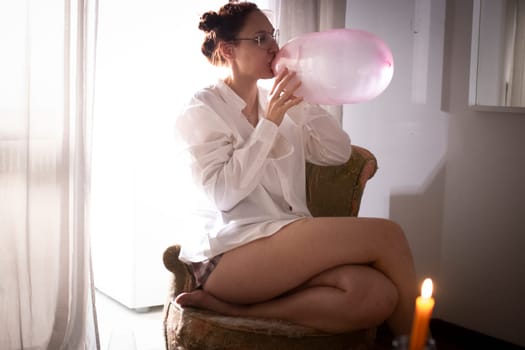 Woman inflating a big pink bubblegum bubble while sitting on a vintage armchair at home