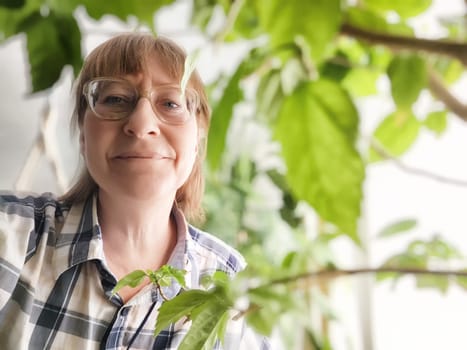 Middle-aged woman with blonde hair smiles amid vibrant green plants at home, embodying tranquility and connection.