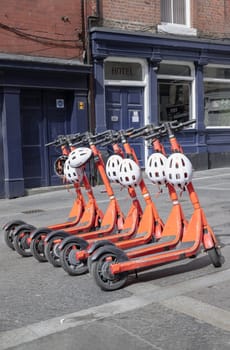Newcastle, UK - Apr 14, 2025 - Bright orange electric scooters with white safety helmets parked on the sidewalk in downtown. Public rental e-scooter for trip and travel in center city, use it as your Wallpaper, Poster and Space for text, Selective focus.