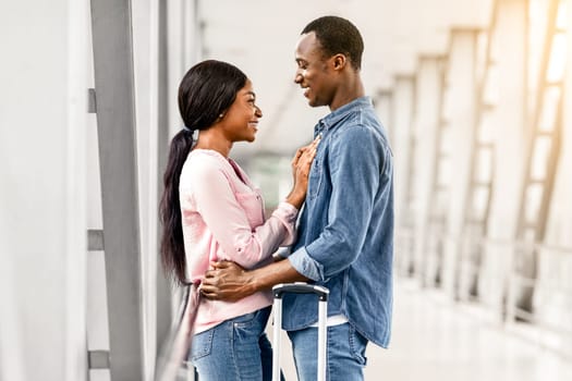 Loving African American Man And Woman Cuddling In Airport Terminal Before Flight, Happy Romantic Black Couple Waiting For Plane Boarding, Ready For Honeymoon Vacation Trip, Side View With Free Space