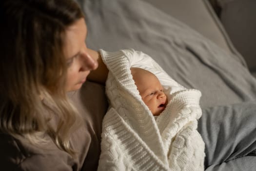 In a calm indoor setting, a mother holds her wrapped newborn, both embracing a peaceful moment of bonding on a soft bed at dusk.