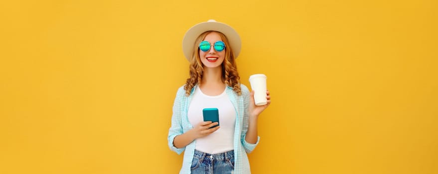 Summer stylish happy young woman with phone, cool girl using smartphone holding cup of coffee dressed tourist straw hat standing on yellow background