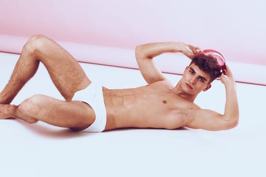 Confident young man in swimwear lying on his side, holding a swimming cap, against a soft pink background. This image represents fitness, summer, and leisure.