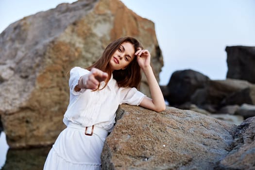 Serene woman in flowing white dress poses confidently on seaside rock with hands on hips