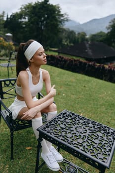 A young female athlete in white sportswear sits in a garden, reflecting a calm demeanor that emphasizes a healthy lifestyle and fitness motivation in a tranquil setting