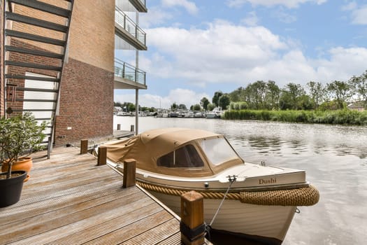 A peaceful riverfront view featuring a small boat moored to a wooden dock, surrounded by greenery and under a clear sky with fluffy clouds.