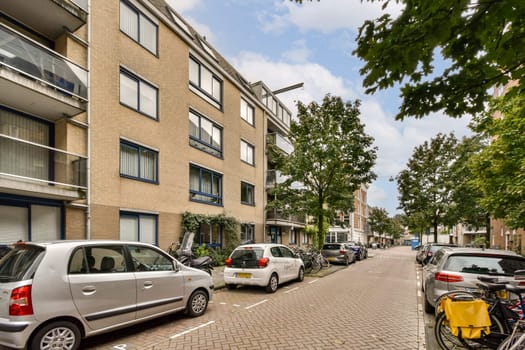 A view of a residential street featuring contemporary apartment buildings and parked cars amidst trees lining the road.