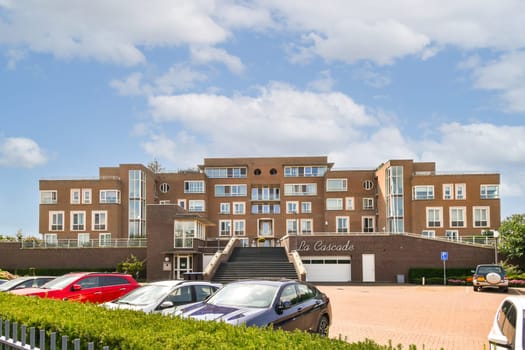 A contemporary apartment building features large windows and balconies, surrounded by greenery and parked cars, set against a bright blue sky.