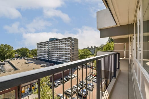 A spacious balcony view showcases modern buildings and a bustling parking lot, framed by trees under a clear blue sky.