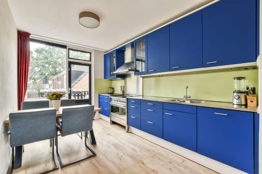 A contemporary kitchen featuring sleek blue cabinets, a tidy dining area, and large windows that offer natural light and views of greenery outside.