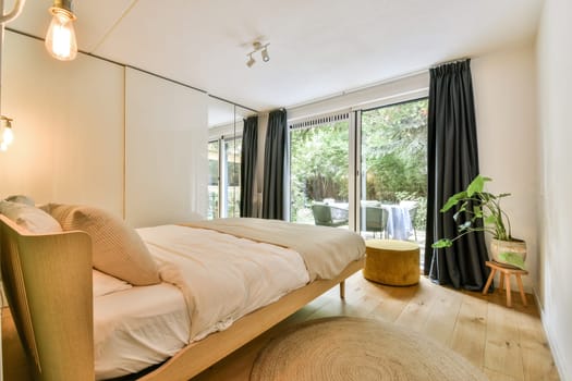 A spacious and bright bedroom featuring minimalist decor, large windows with greenery outside, and cozy furnishings creating a serene atmosphere.