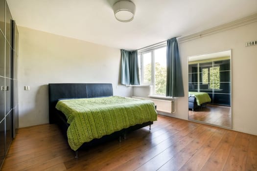 A spacious modern bedroom featuring a bed with a green patterned bedspread, a mirror, and large windows allowing natural light.
