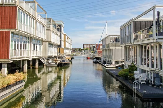 Stylish modern houses line a serene waterway, reflecting a blend of architecture and nature. Boats are moored along the canal, enhancing the picturesque setting.