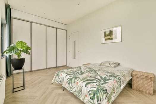 A stylish minimalist bedroom featuring a floral tropical patterned bedspread, large windows, and a decorative potted plant, emphasizing comfort and elegance.