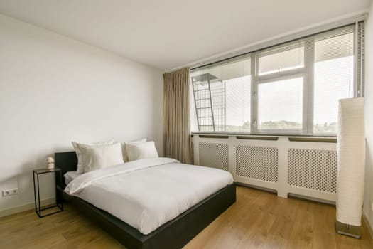A spacious, modern bedroom featuring a neatly made bed, large windows with sheer curtains, and minimal decor. The warm wooden floor enhances the serene ambiance.