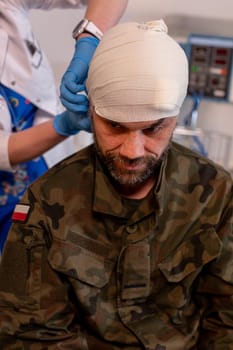 A soldier suffering from a significant head injury is undergoing urgent bandaging in a military medical facility