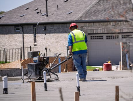 A construction worker uses a concrete finishing machine to smooth a fresh concrete slab on a new foundation