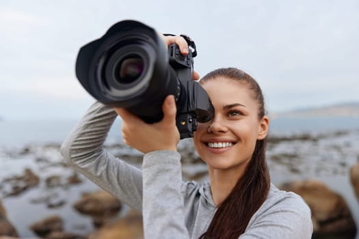 A cheerful young woman with long hair is enjoying photography at the beach, showcasing her passion for capturing vibrant nature moments with a professional camera