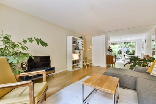A cozy modern living room features green plants, modern furniture, and natural light. The space blends comfort with a stylish decor, ideal for relaxing or socializing.